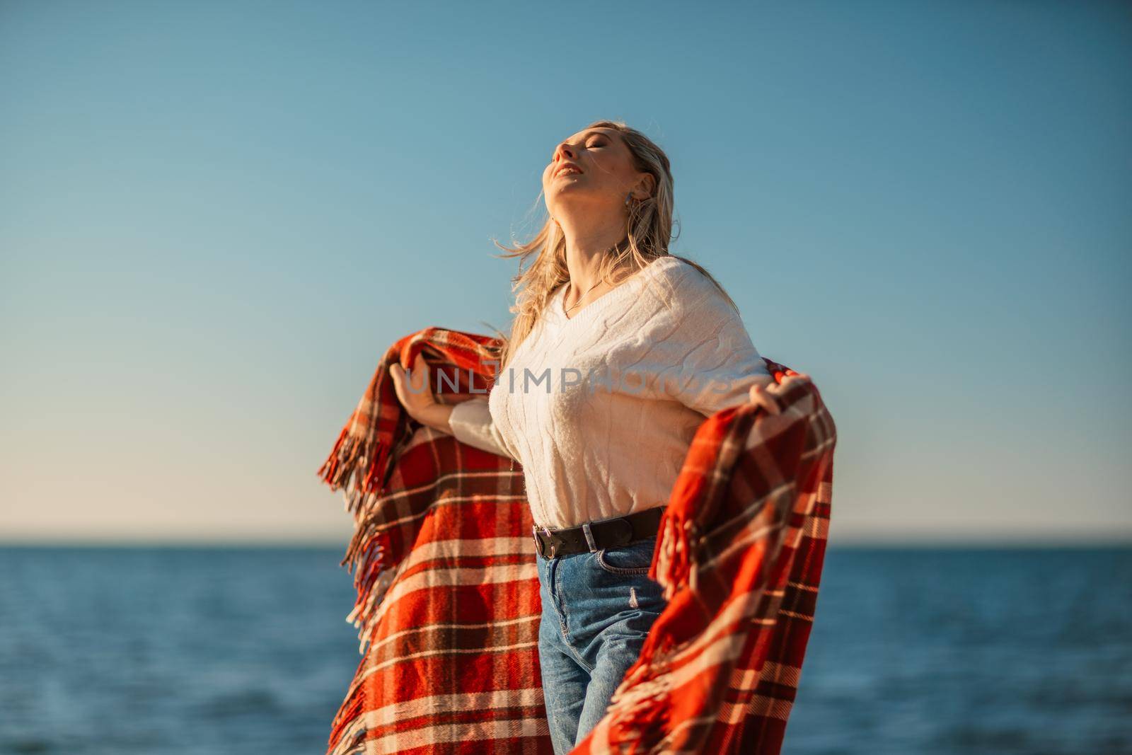 Attractive blonde Caucasian woman enjoying time on the beach at sunset, walking in a blanket and looking to the side, with the sunset sky and sea in the background. Beach vacation.