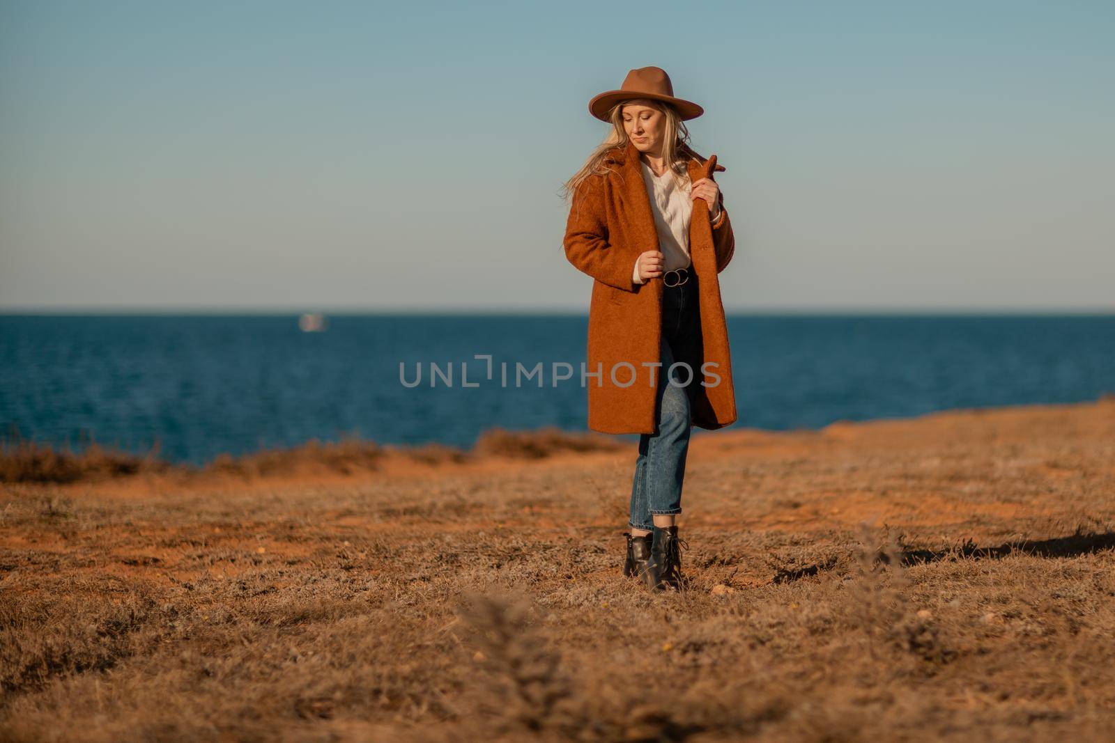 A woman walking along the coast near the sea. An elegant lady in a brown coat and a hat with fashionable makeup walks on the seashore.