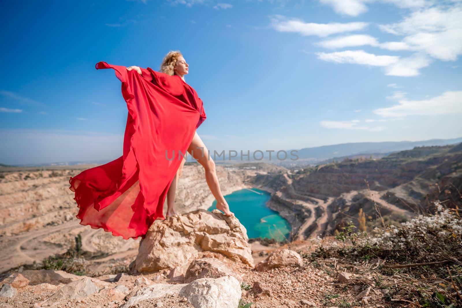 Side view of a beautiful sensual woman in a red long dress posing on a rock high above the lake in the afternoon. Against the background of the blue sky and the lake in the form of a heart by Matiunina