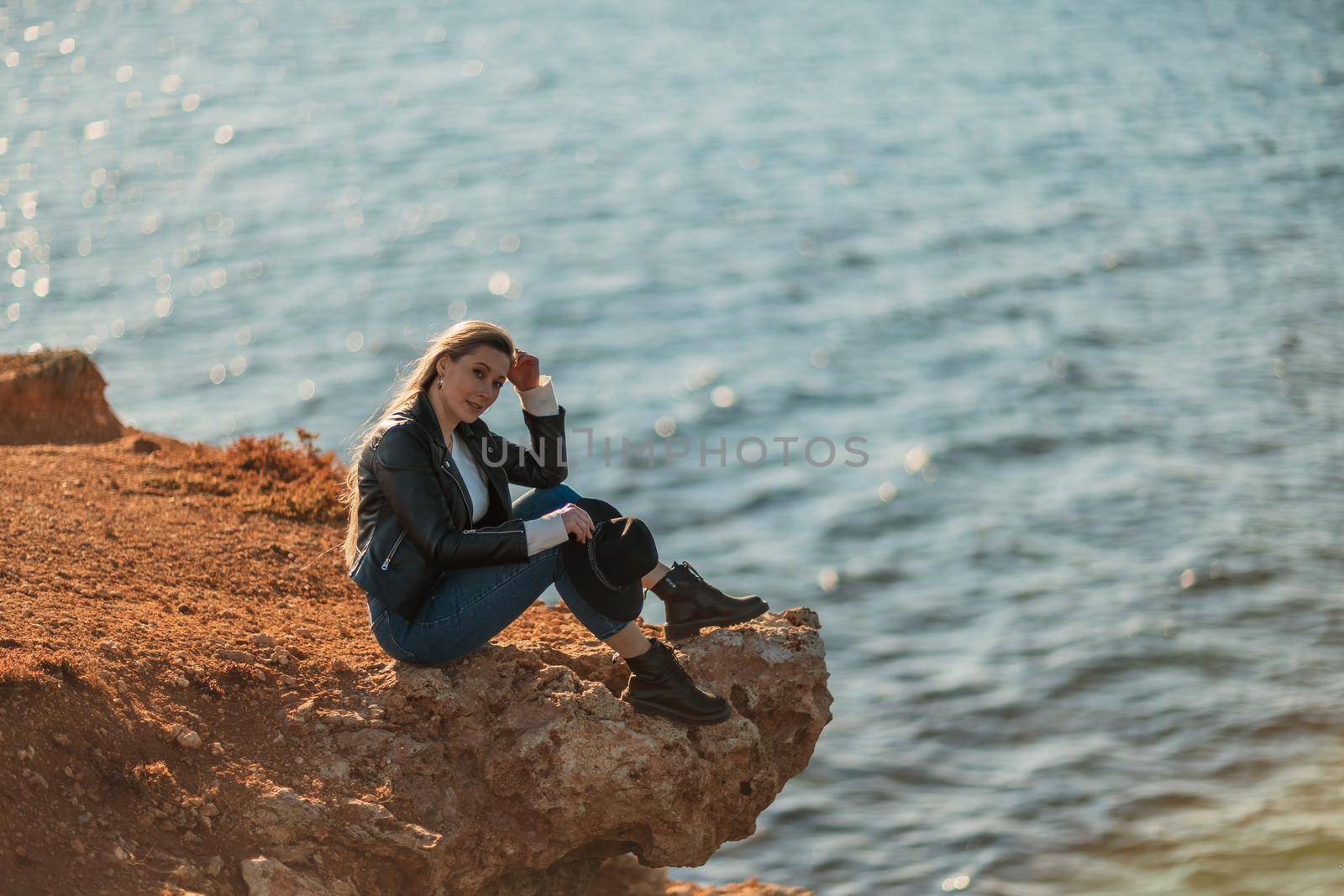 A blonde girl in a stylish black leather jacket is sitting on the seashore