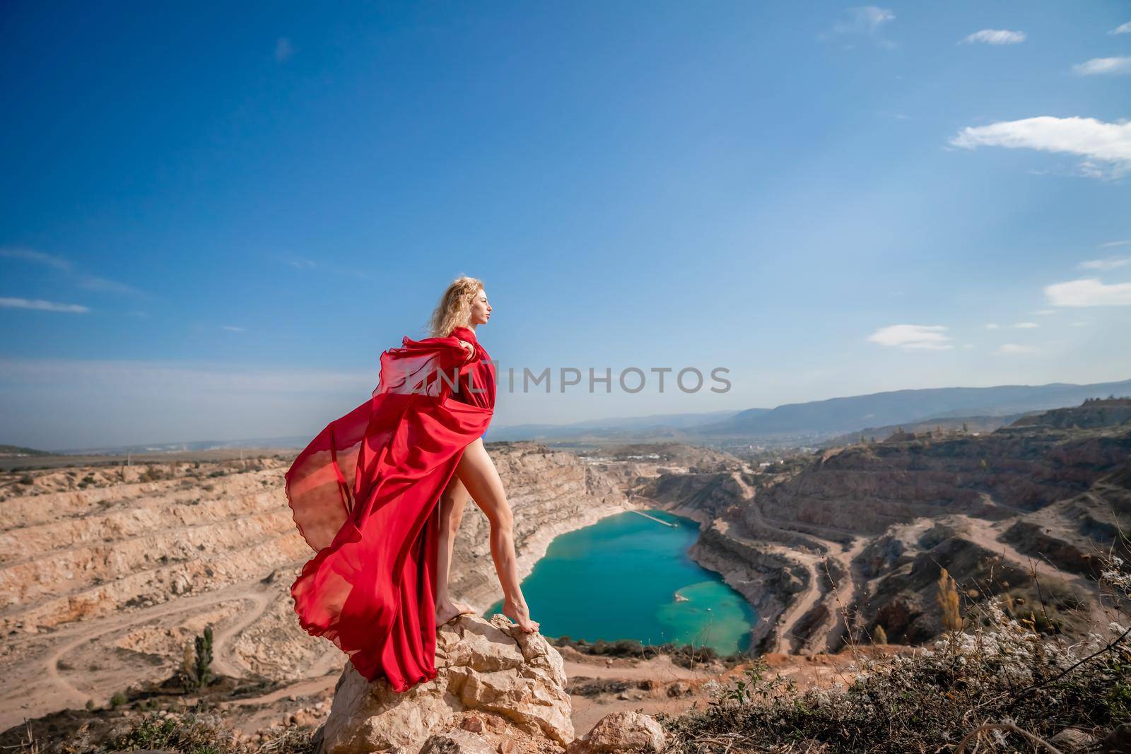 Side view of a beautiful sensual woman in a red long dress posing on a rock high above the lake in the afternoon. Against the background of the blue sky and the lake in the form of a heart by Matiunina