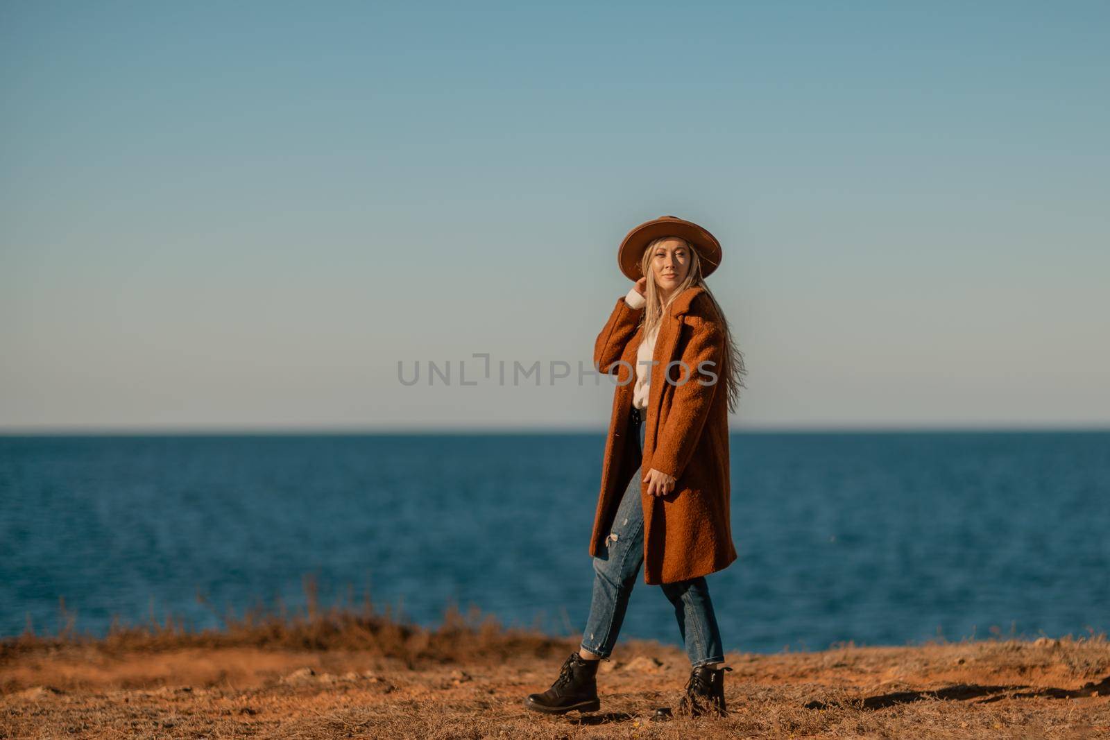 A woman walking along the coast near the sea. An elegant lady in a brown coat and a hat with fashionable makeup walks on the seashore by Matiunina