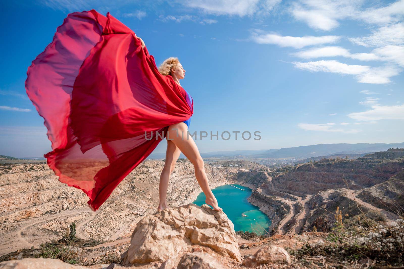 Side view of a beautiful sensual woman in a red long dress posing on a rock high above the lake in the afternoon. Against the background of the blue sky and the lake in the form of a heart.