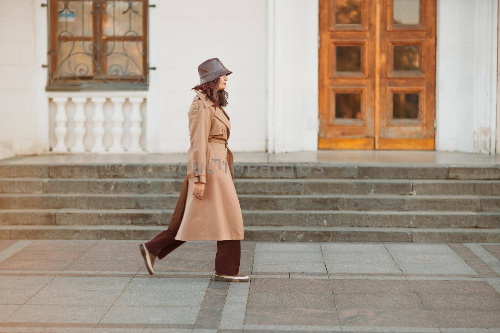 Outdoor fashion portrait of young elegant fashionable brunette woman, model in stylish hat, choker and light raincoat posing at sunset in European city. by Matiunina