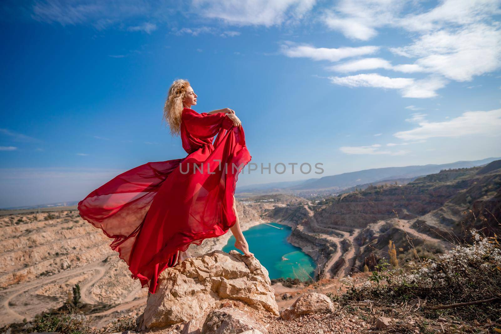 Side view of a beautiful sensual woman in a red long dress posing on a rock high above the lake in the afternoon. Against the background of the blue sky and the lake in the form of a heart.