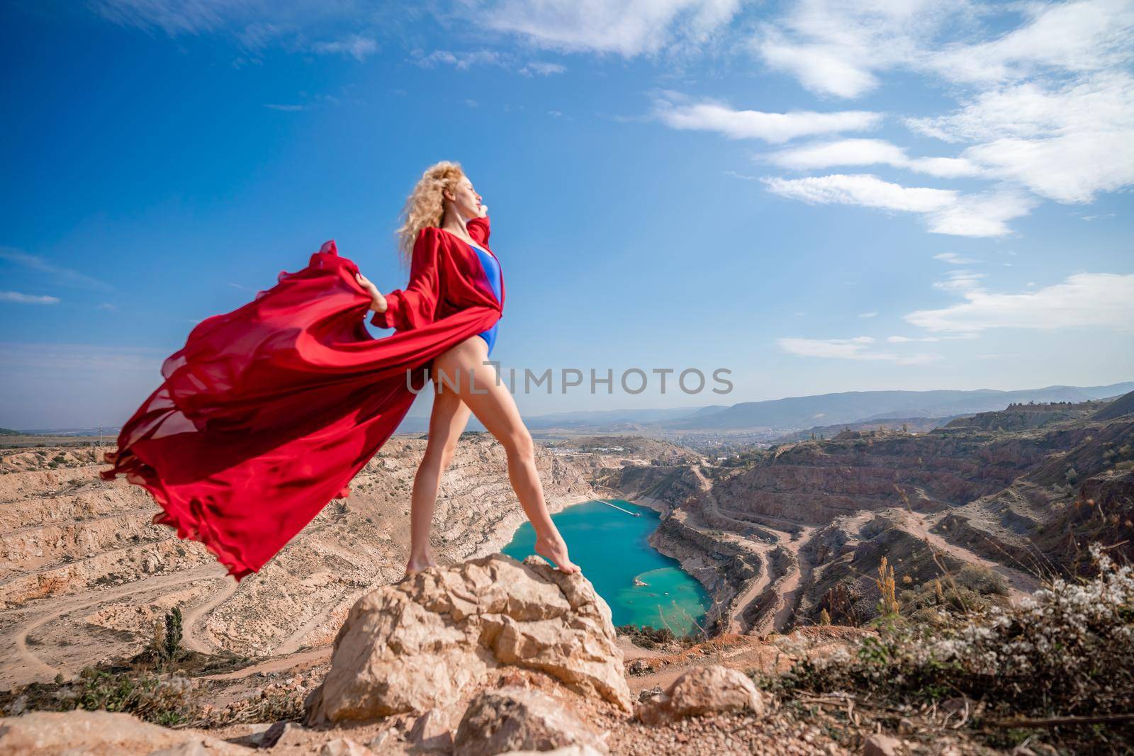 Side view of a beautiful sensual woman in a red long dress posing on a rock high above the lake in the afternoon. Against the background of the blue sky and the lake in the form of a heart by Matiunina