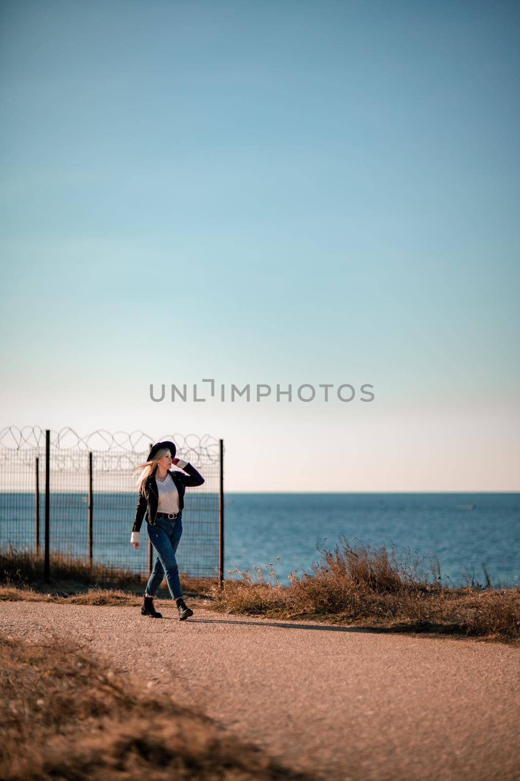 A blonde in a stylish black leather jacket walks along the seashore. by Matiunina