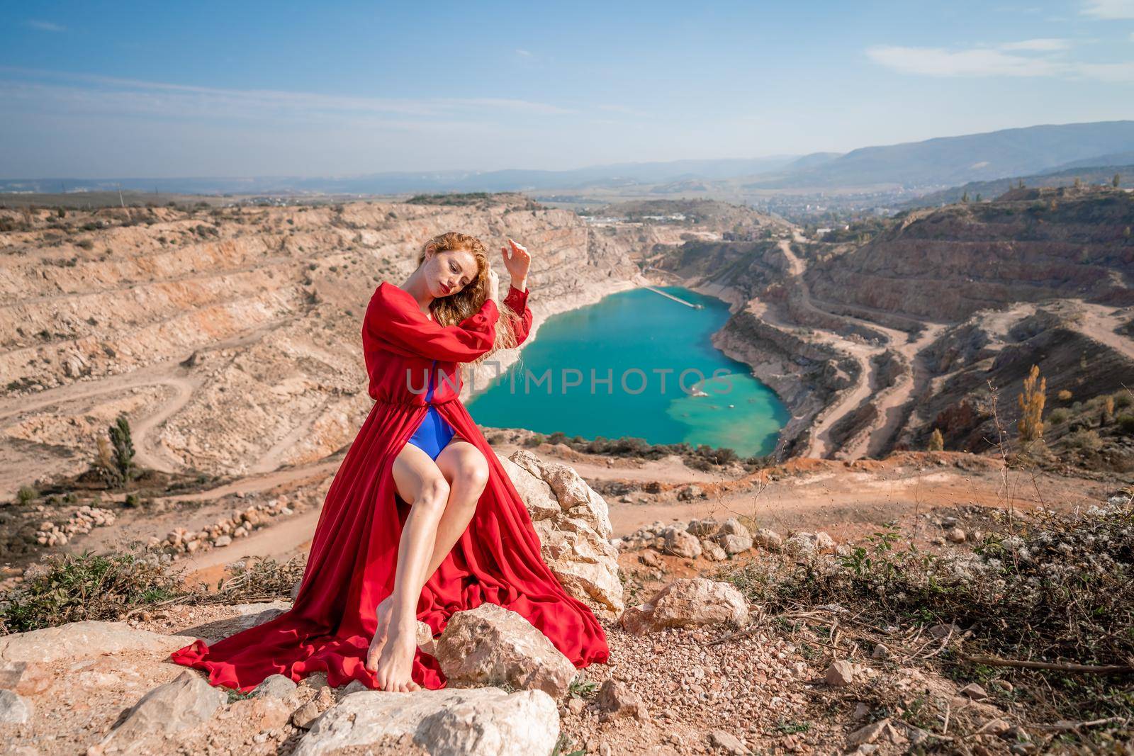 A beautiful girl in a red long dress, Sits on a rock high above the lake in the afternoon. Against the background of the blue sky and the lake in the shape of a heart. by Matiunina