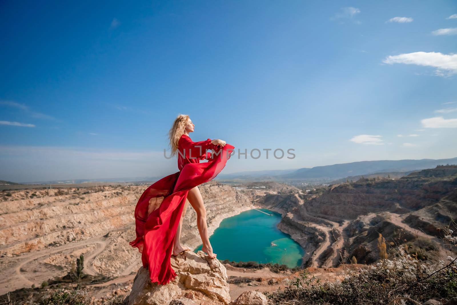 Side view of a beautiful sensual woman in a red long dress posing on a rock high above the lake in the afternoon. Against the background of the blue sky and the lake in the form of a heart by Matiunina