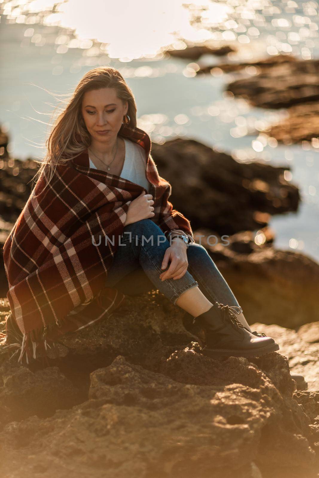Attractive blonde Caucasian woman enjoying time on the beach at sunset, sitting in a blanket and looking to the side, with the sunset sky and sea in the background. Beach vacation. by Matiunina