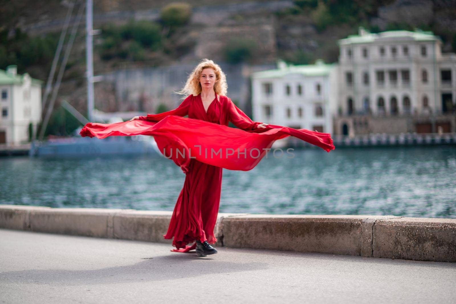 A woman in a red dress, a fashion model with long silk wings in a flowing dress, flying fabric on the embankment. by Matiunina