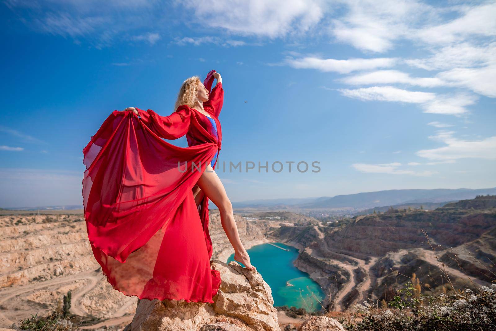 Side view of a beautiful sensual woman in a red long dress posing on a rock high above the lake in the afternoon. Against the background of the blue sky and the lake in the form of a heart.