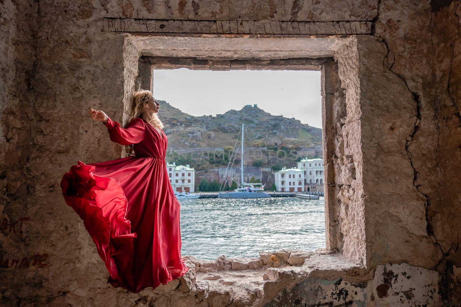 View of Balaklava Bay through an arched balcony in oriental style. The girl in a long red dress stands with her back. Abandoned mansion on the Black Sea coast.