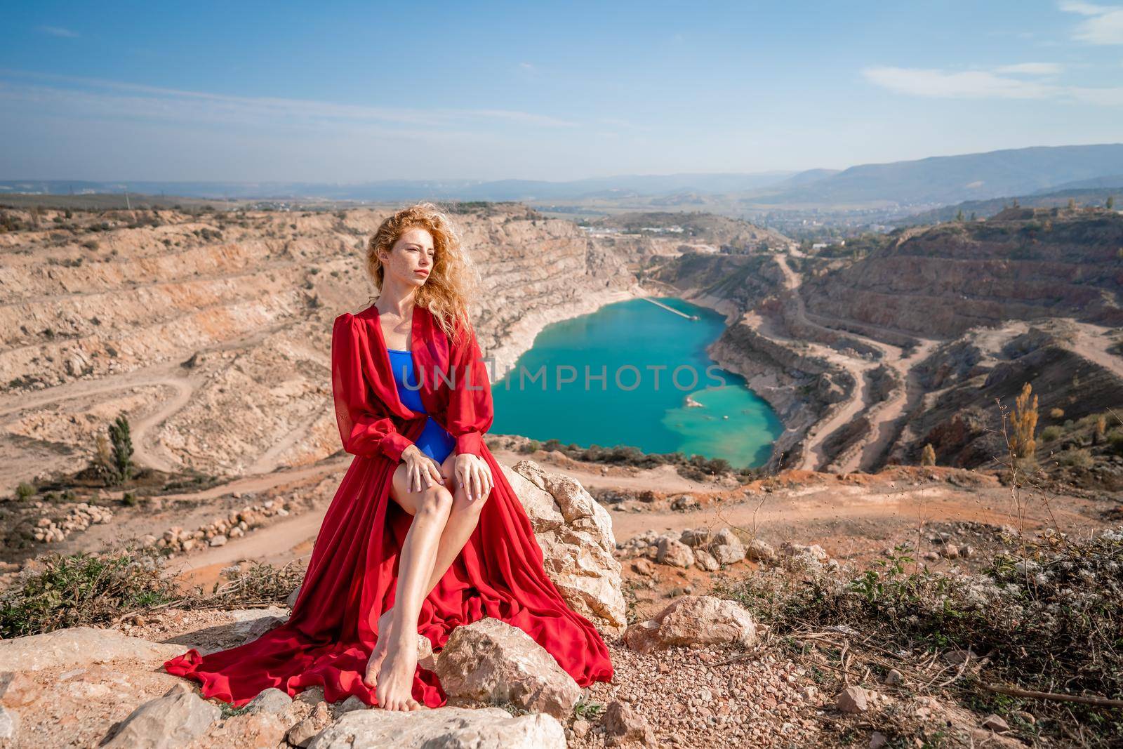 A beautiful girl in a red long dress, Sits on a rock high above the lake in the afternoon. Against the background of the blue sky and the lake in the shape of a heart. by Matiunina