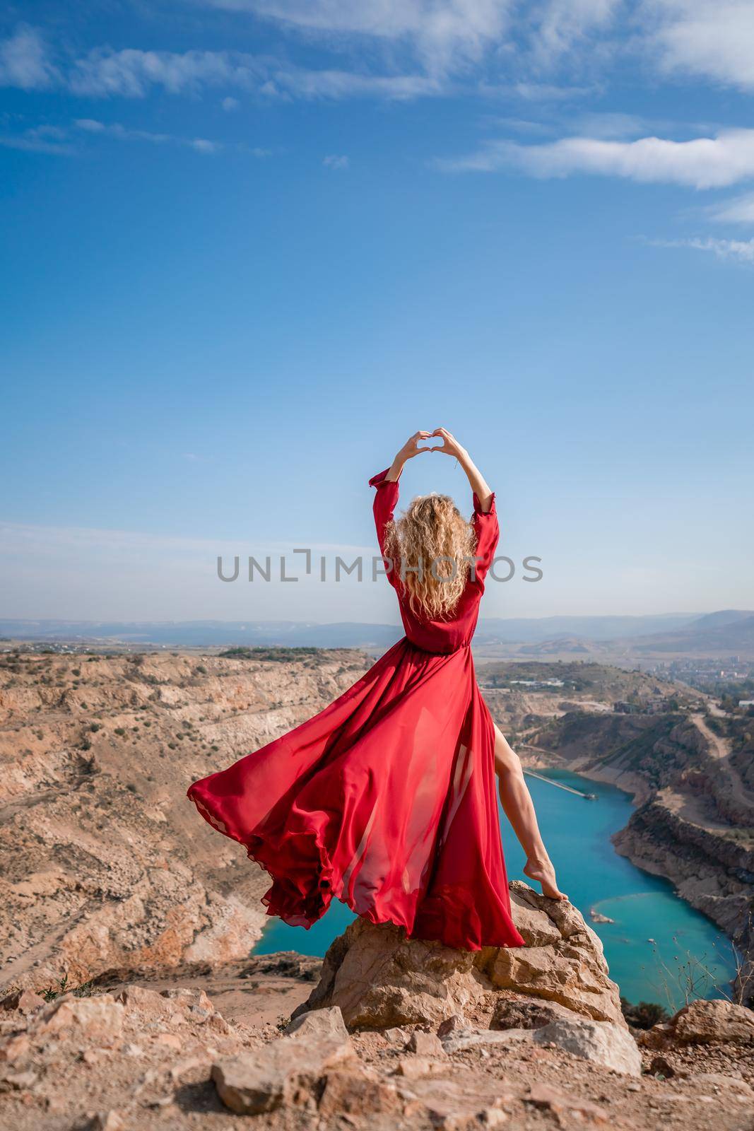 Rear view of a beautiful sensual woman in a red long dress posing on a rock high above the lake in the afternoon. Against the background of the blue sky and the lake in the shape of a heart.