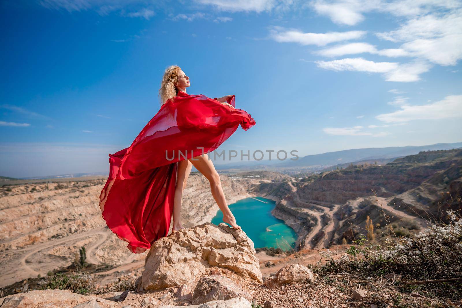 Side view of a beautiful sensual woman in a red long dress posing on a rock high above the lake in the afternoon. Against the background of the blue sky and the lake in the form of a heart.