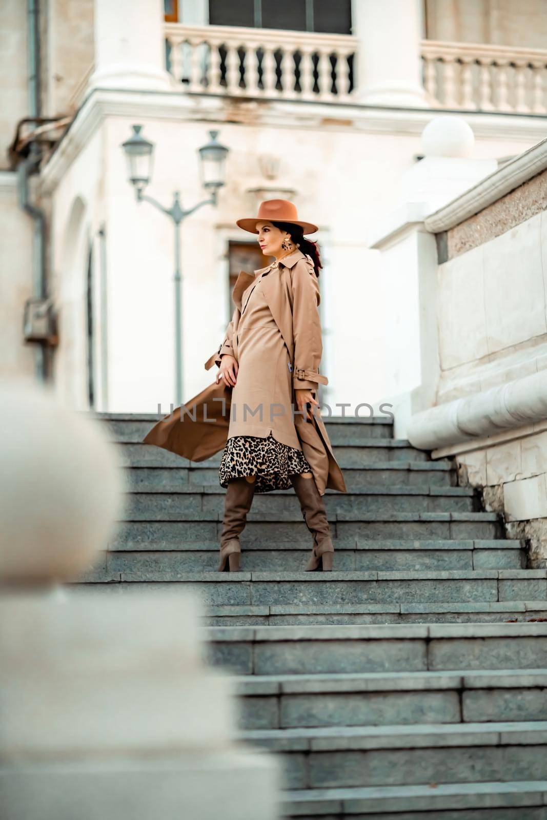 Outdoor fashion portrait of young elegant fashionable brunette woman, model in stylish hat, choker and light raincoat posing at sunset in European city