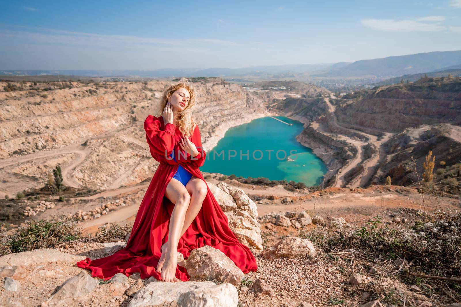 A beautiful girl in a red long dress, Sits on a rock high above the lake in the afternoon. Against the background of the blue sky and the lake in the shape of a heart