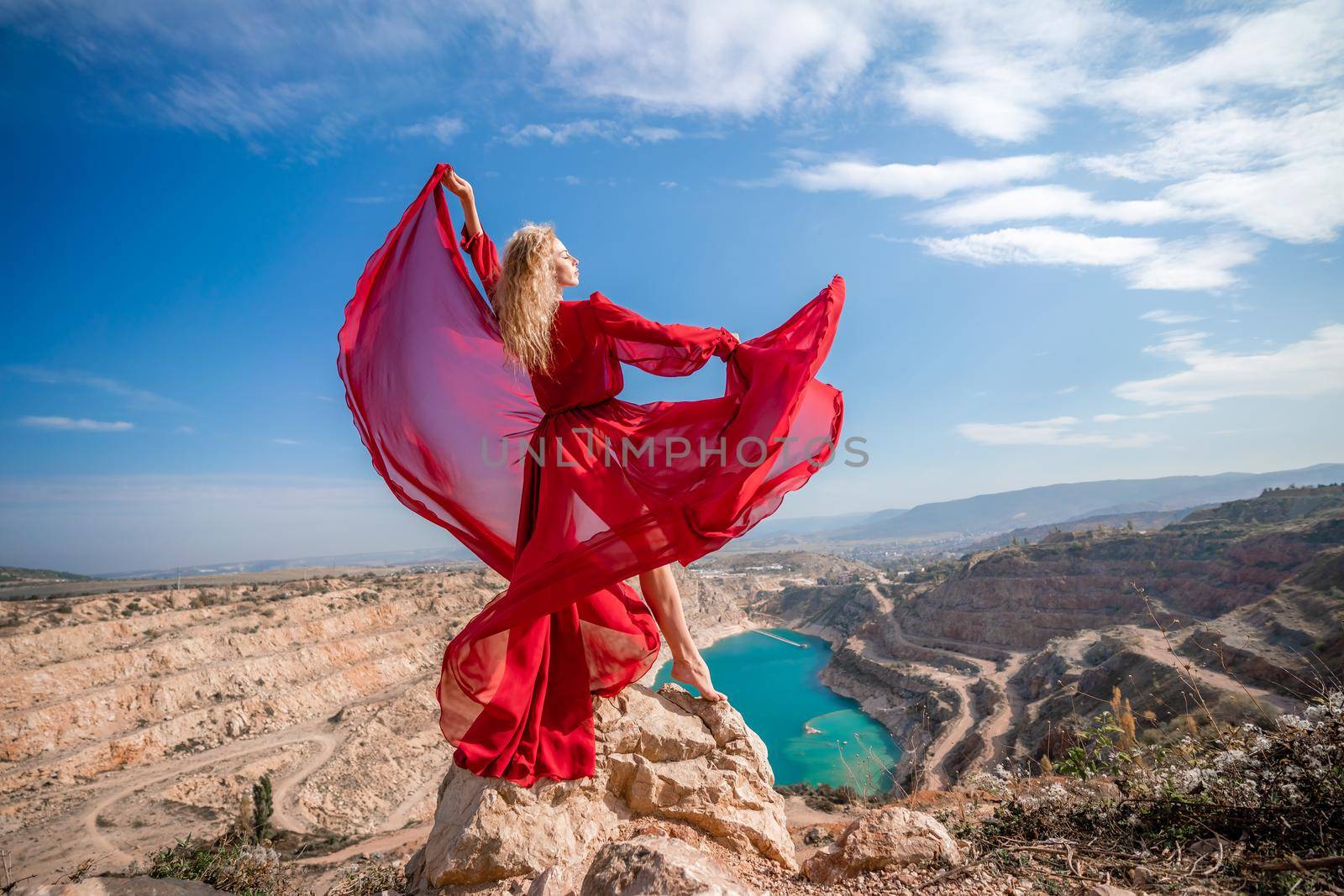 Side view of a beautiful sensual woman in a red long dress posing on a rock high above the lake in the afternoon. Against the background of the blue sky and the lake in the form of a heart by Matiunina