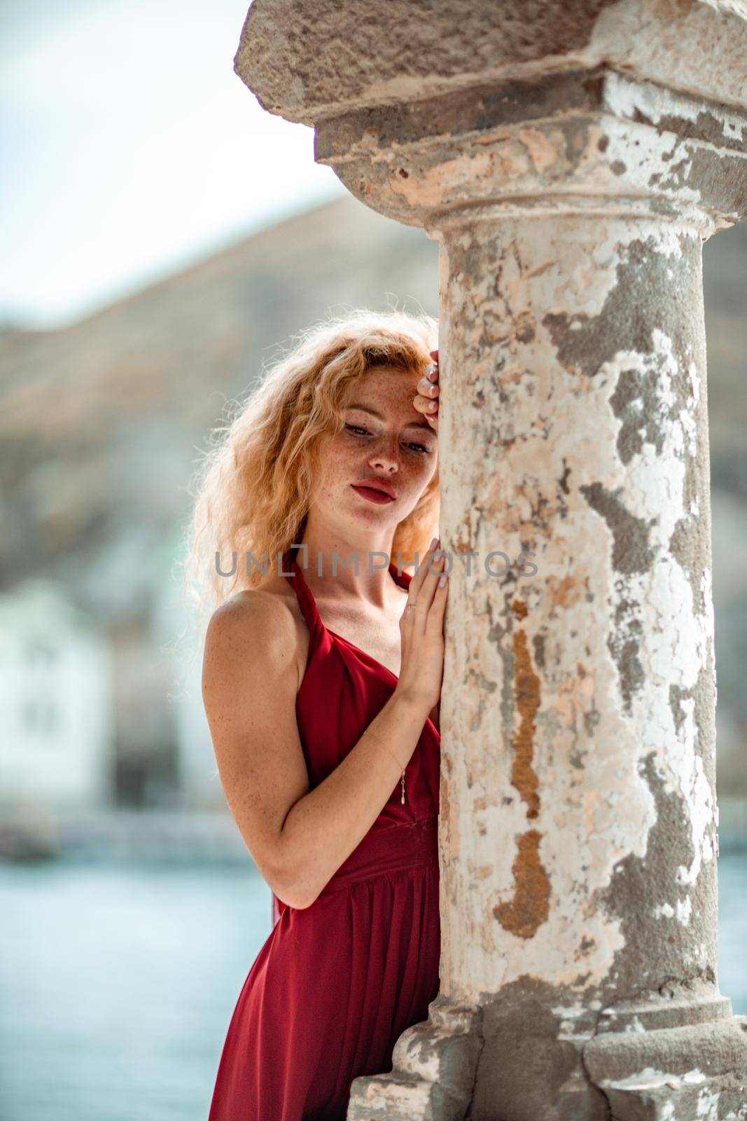 Outdoor portrait of a young beautiful natural redhead girl with freckles, long curly hair, in a red dress, posing against the background of the sea. by Matiunina