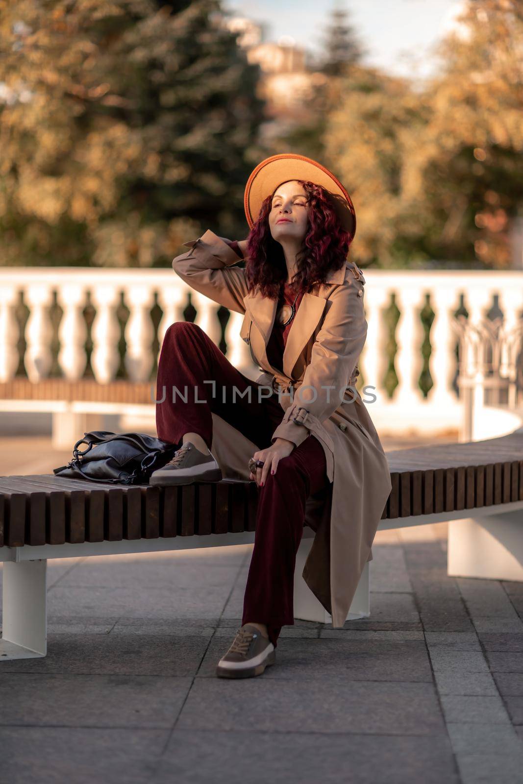 Outdoor fashion portrait of young elegant fashionable brunette woman, model in stylish hat, choker and light raincoat posing at sunset in European city