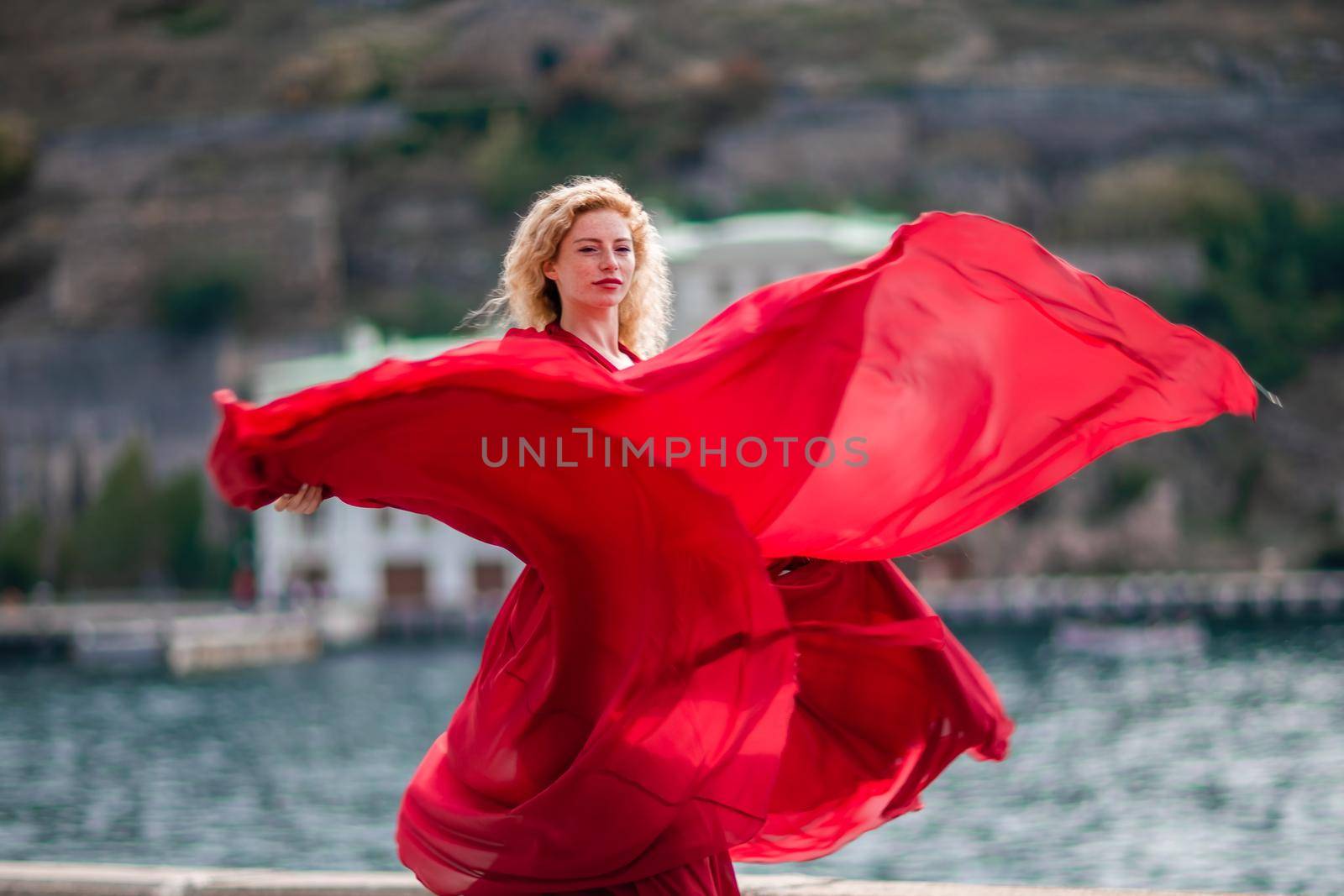 A woman in a red dress, a fashion model with long silk wings in a flowing dress, flying fabric on the embankment