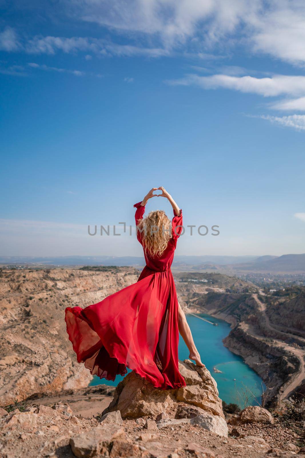 Rear view of a beautiful sensual woman in a red long dress posing on a rock high above the lake in the afternoon. Against the background of the blue sky and the lake in the shape of a heart by Matiunina