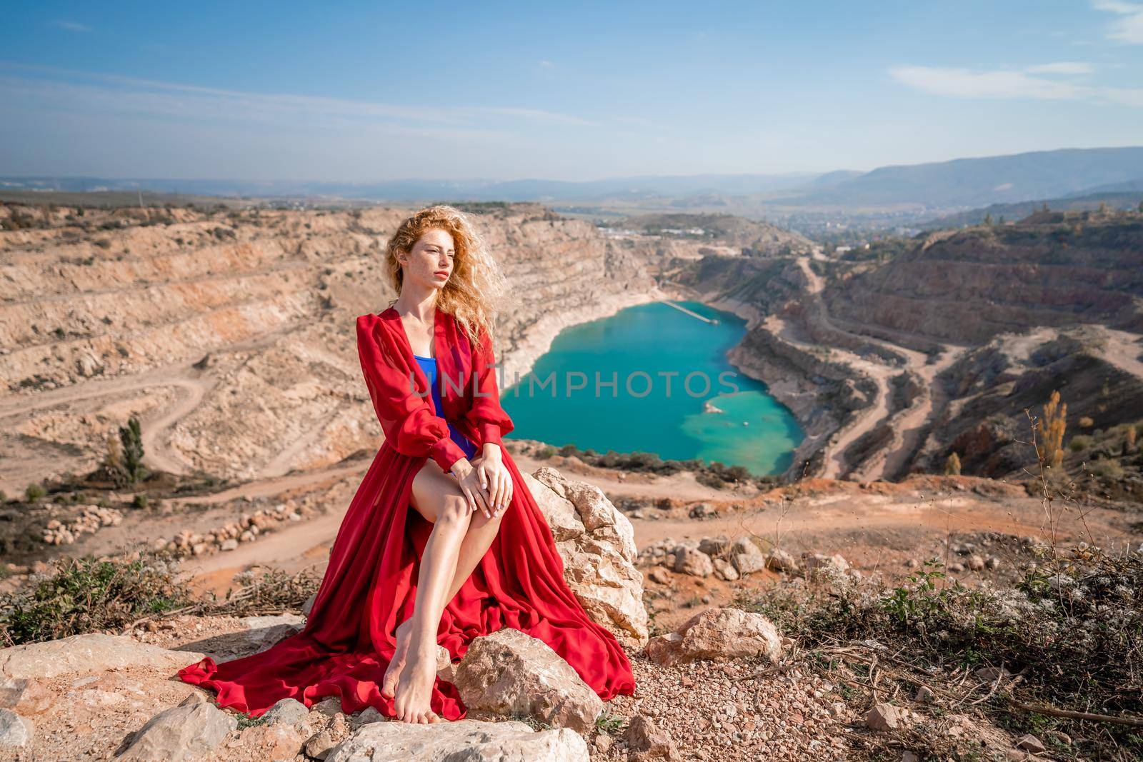 A beautiful girl in a red long dress, Sits on a rock high above the lake in the afternoon. Against the background of the blue sky and the lake in the shape of a heart