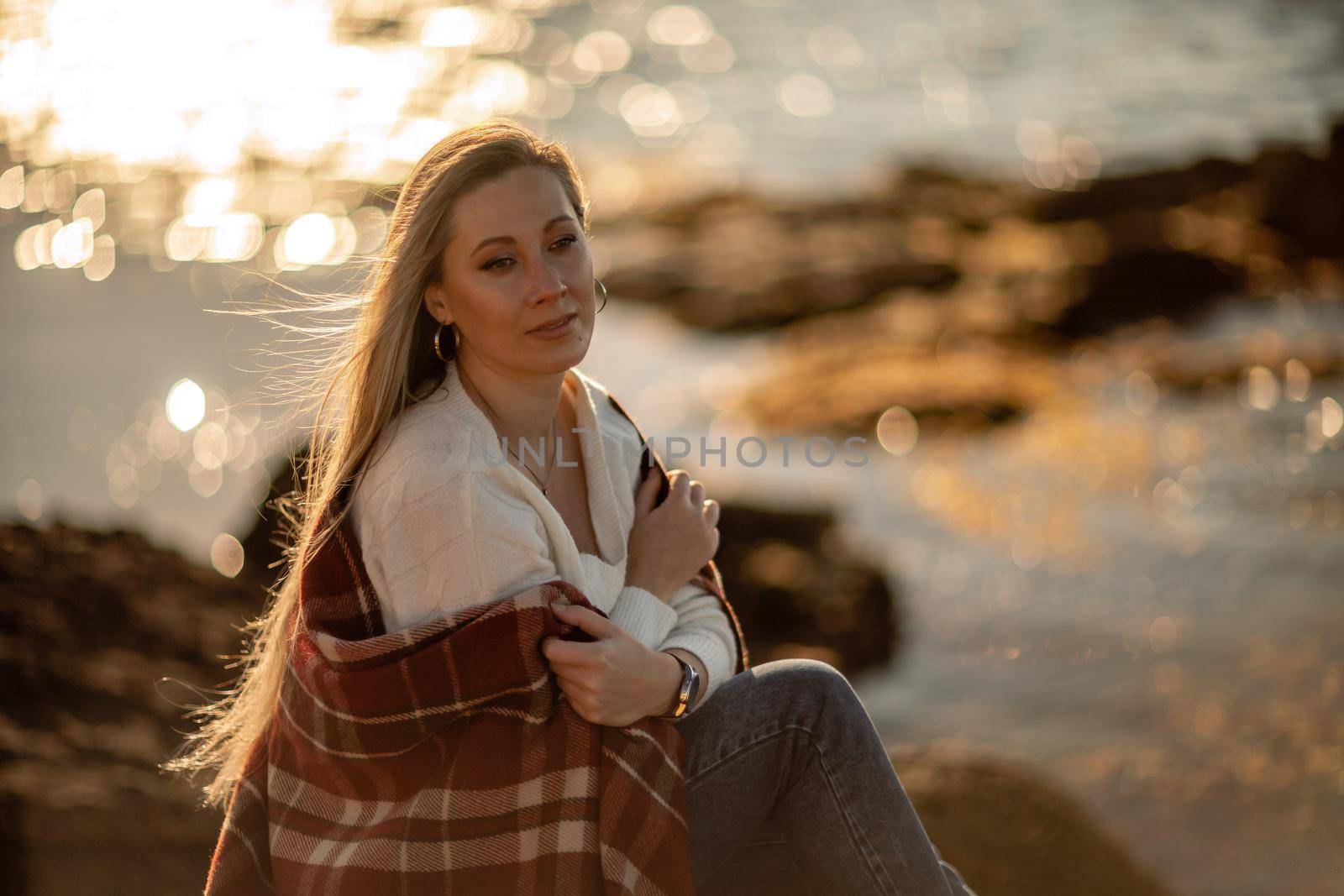 Attractive blonde Caucasian woman enjoying time on the beach at sunset, sitting in a blanket and looking to the side, with the sunset sky and sea in the background. Beach vacation