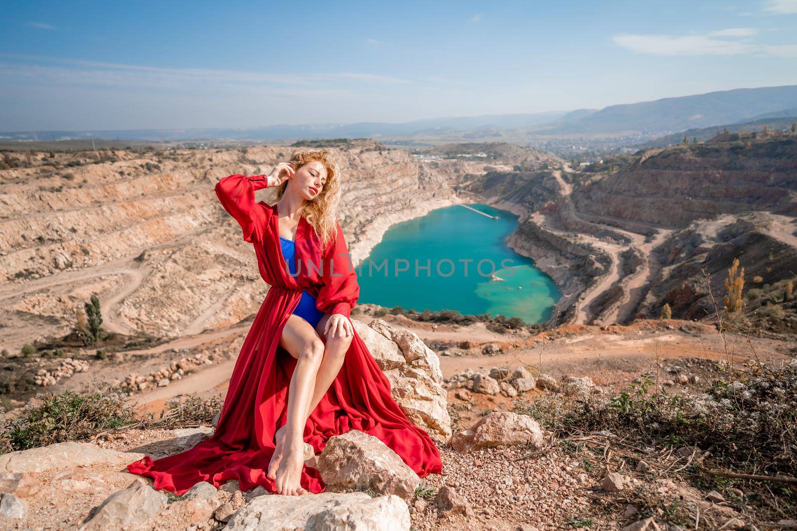 A beautiful girl in a red long dress, Sits on a rock high above the lake in the afternoon. Against the background of the blue sky and the lake in the shape of a heart. by Matiunina