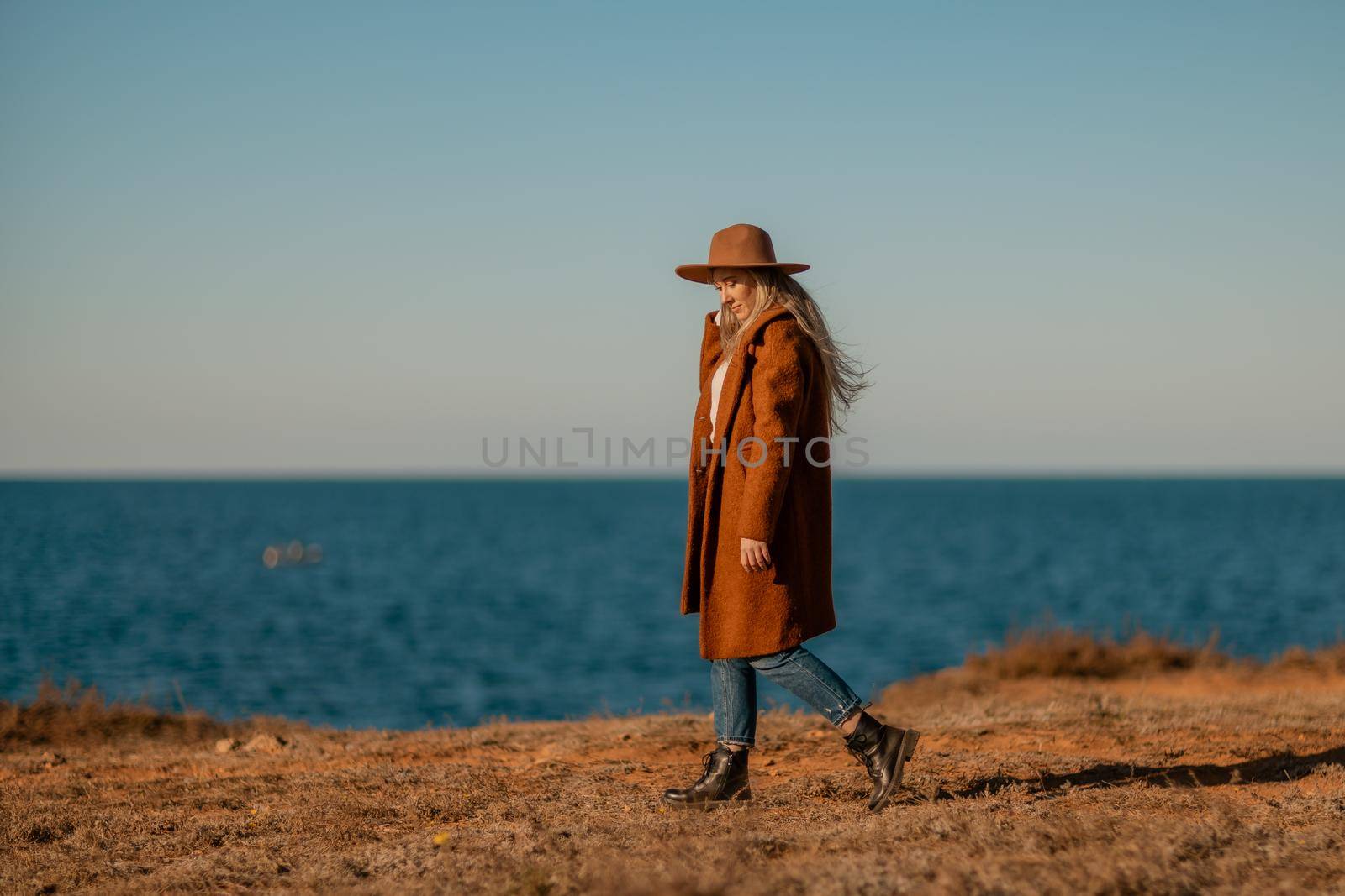 A woman walking along the coast near the sea. An elegant lady in a brown coat and a hat with fashionable makeup walks on the seashore.