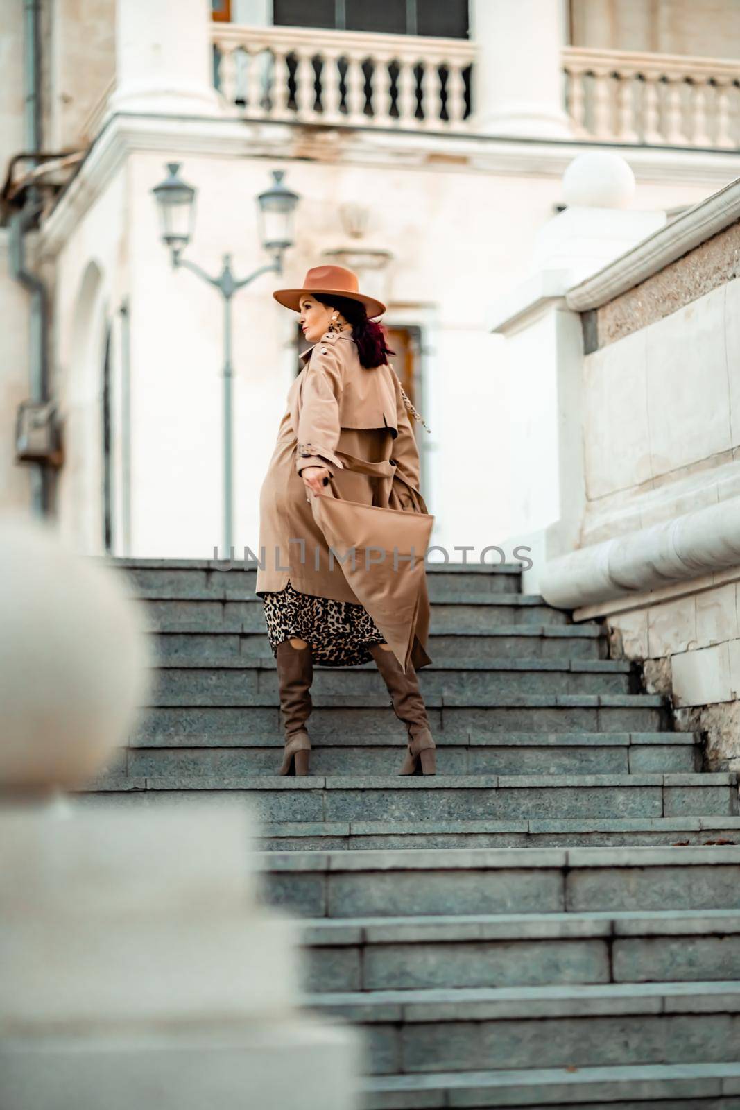 Outdoor fashion portrait of young elegant fashionable brunette woman, model in stylish hat, choker and light raincoat posing at sunset in European city