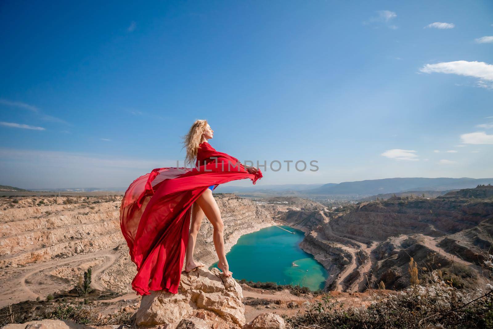 Side view of a beautiful sensual woman in a red long dress posing on a rock high above the lake in the afternoon. Against the background of the blue sky and the lake in the form of a heart.