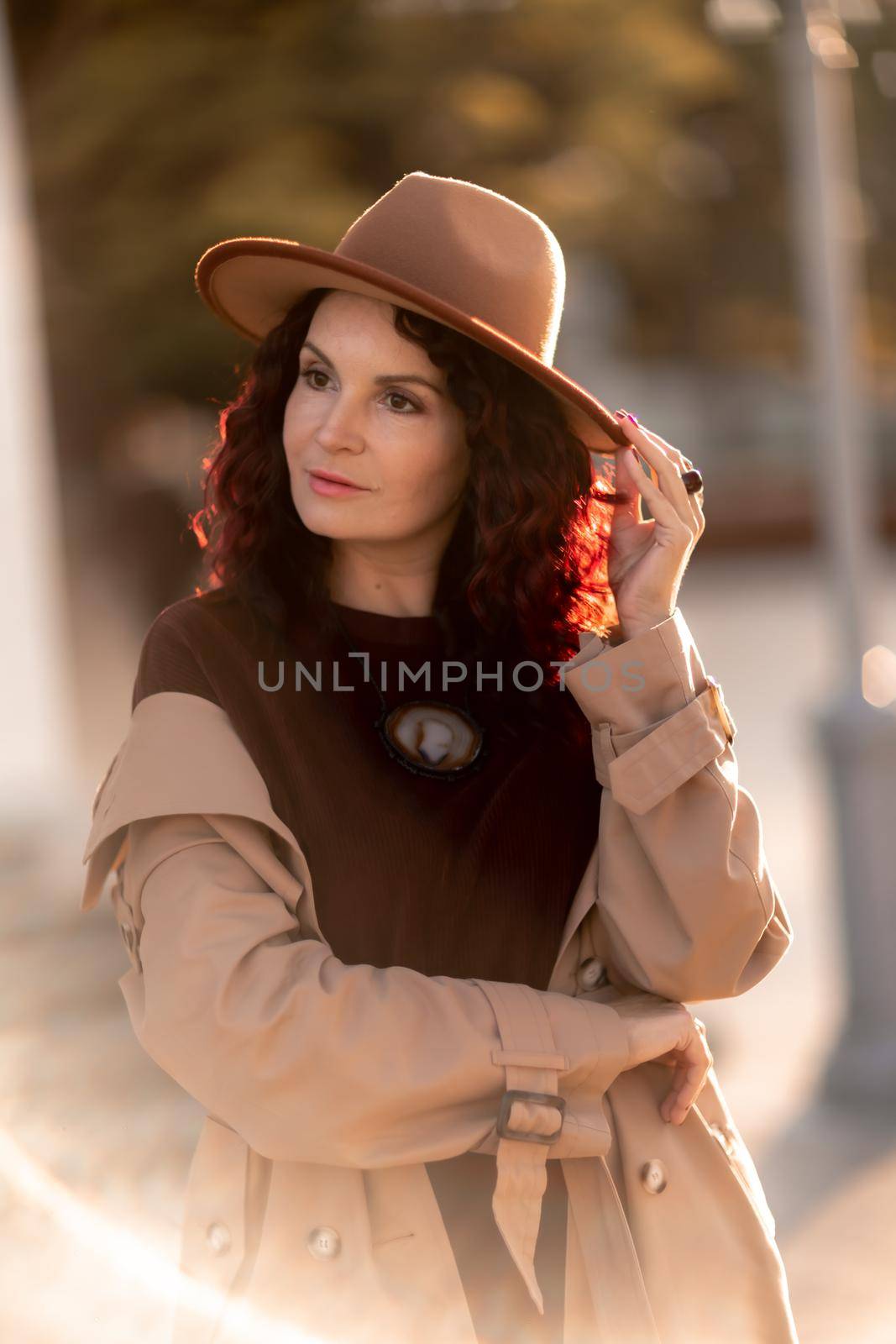 Outdoor fashion portrait of young elegant fashionable brunette woman, model in stylish hat, choker and light raincoat posing at sunset in European city