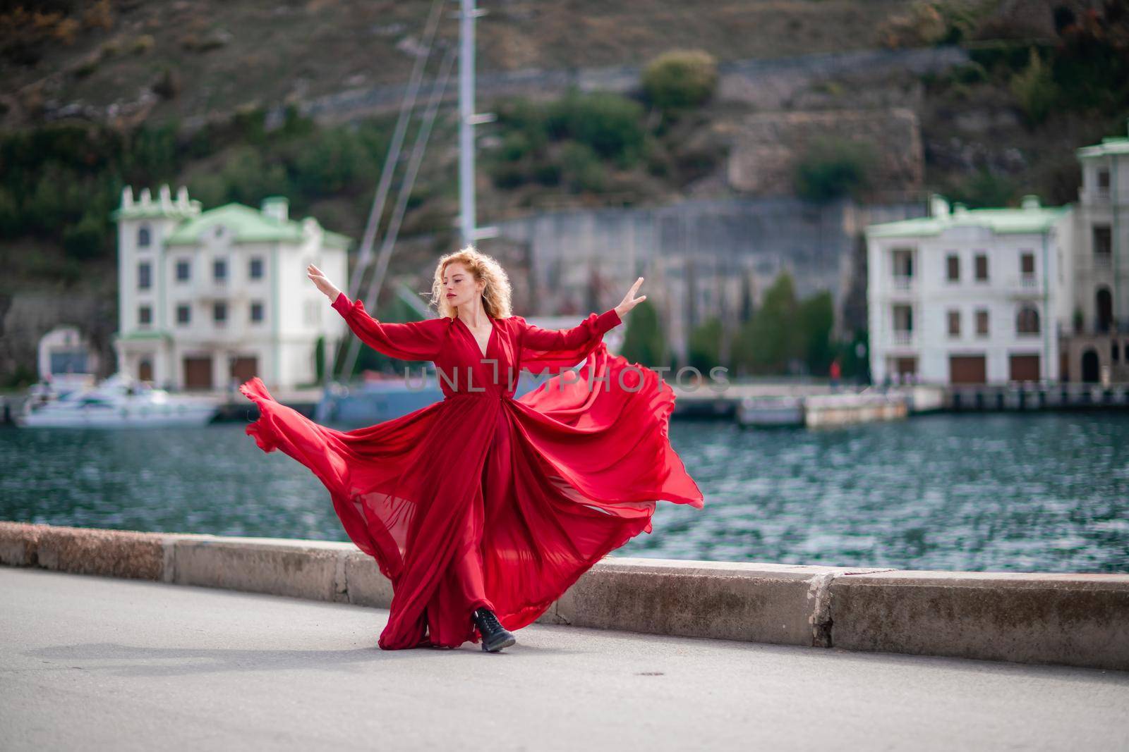 A woman in a red dress, a fashion model with long silk wings in a flowing dress, flying fabric on the embankment