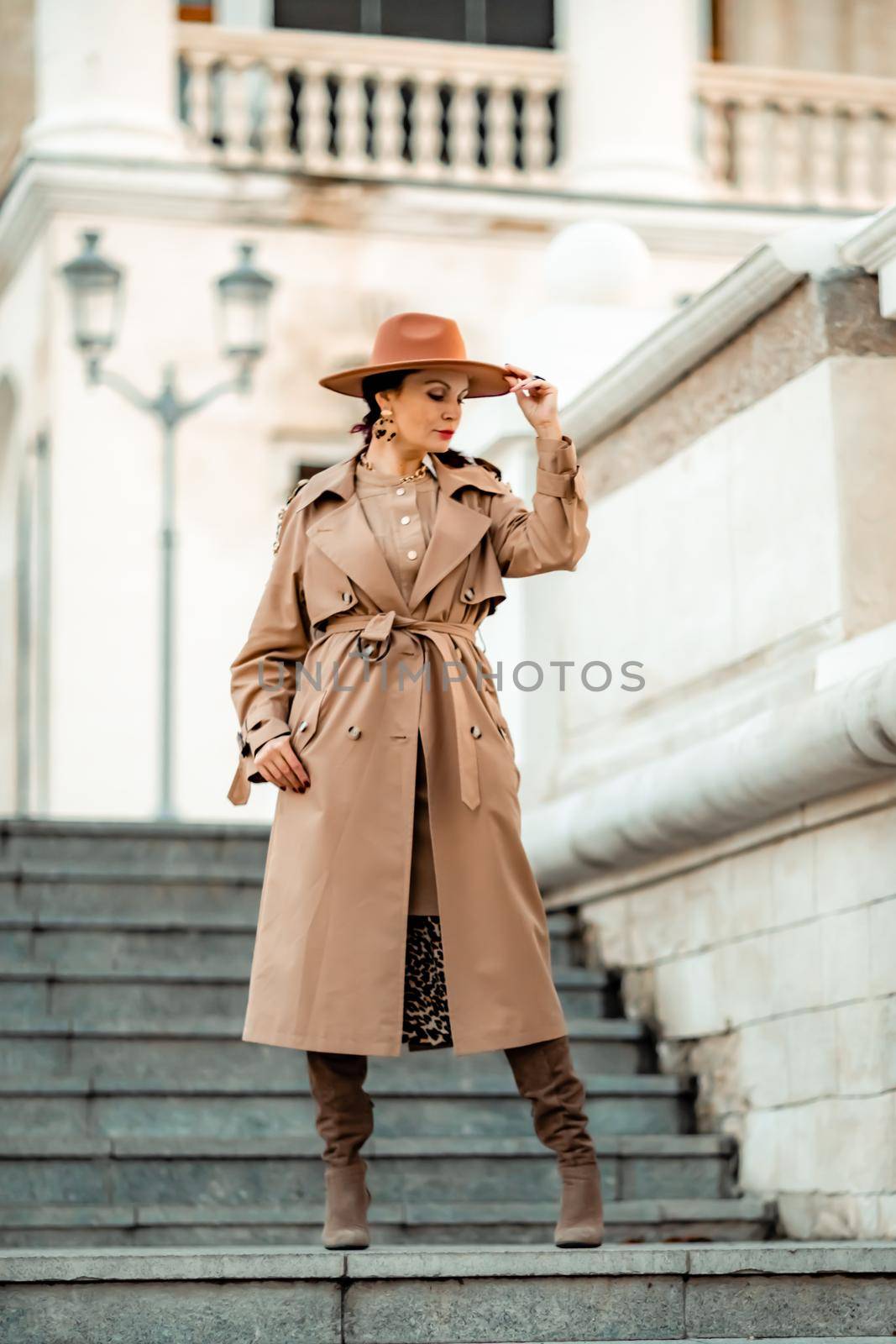 Outdoor fashion portrait of young elegant fashionable brunette woman, model in stylish hat, choker and light raincoat posing at sunset in European city