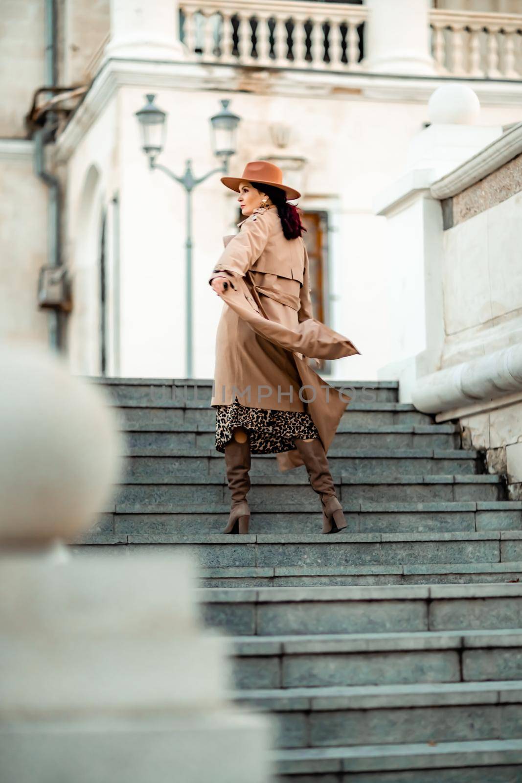 Outdoor fashion portrait of young elegant fashionable brunette woman, model in stylish hat, choker and light raincoat posing at sunset in European city. by Matiunina