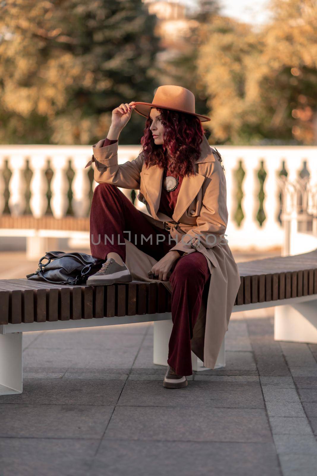 Outdoor fashion portrait of young elegant fashionable brunette woman, model in stylish hat, choker and light raincoat posing at sunset in European city