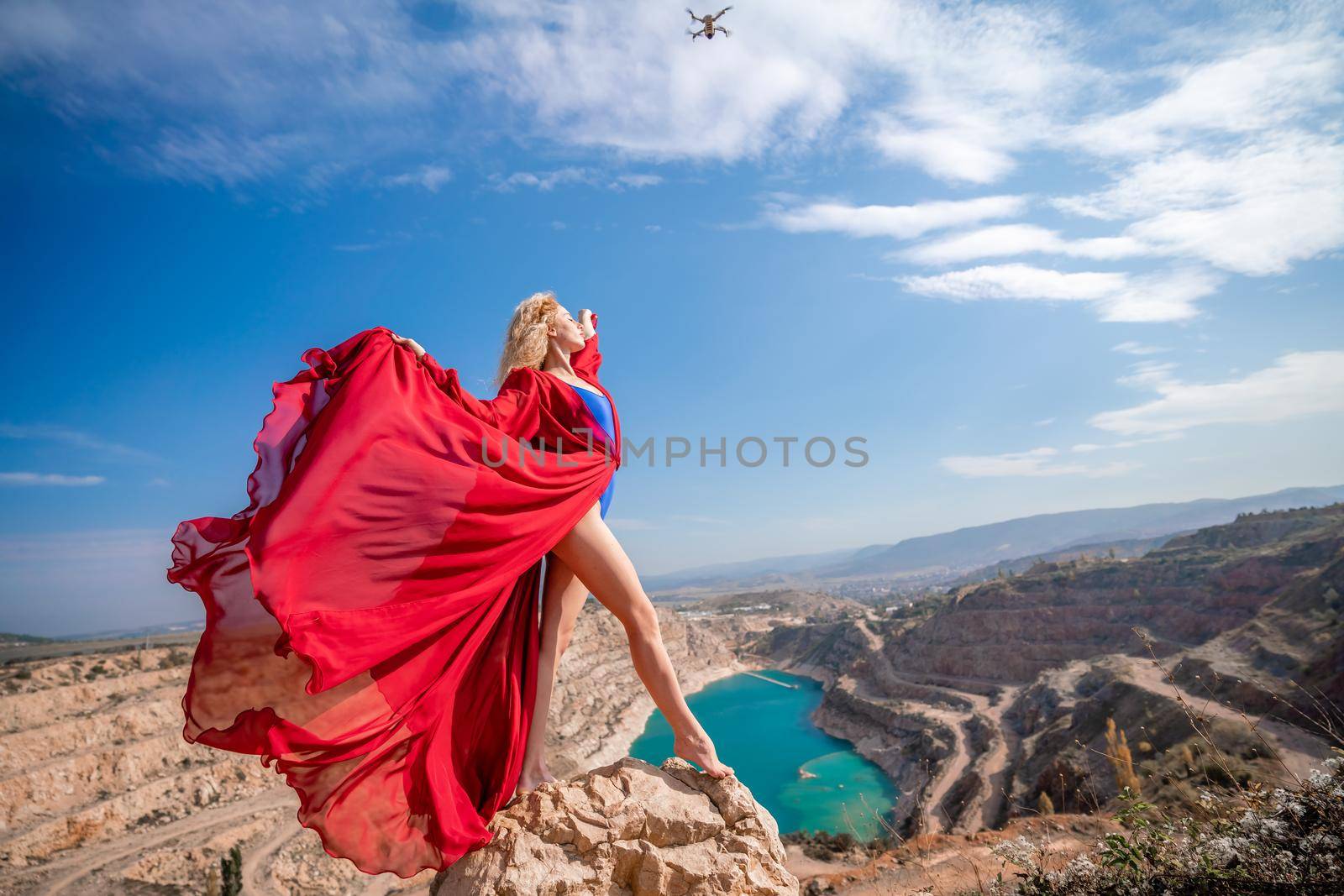 Side view of a beautiful sensual woman in a red long dress posing on a rock high above the lake in the afternoon. Against the background of the blue sky and the lake in the form of a heart.