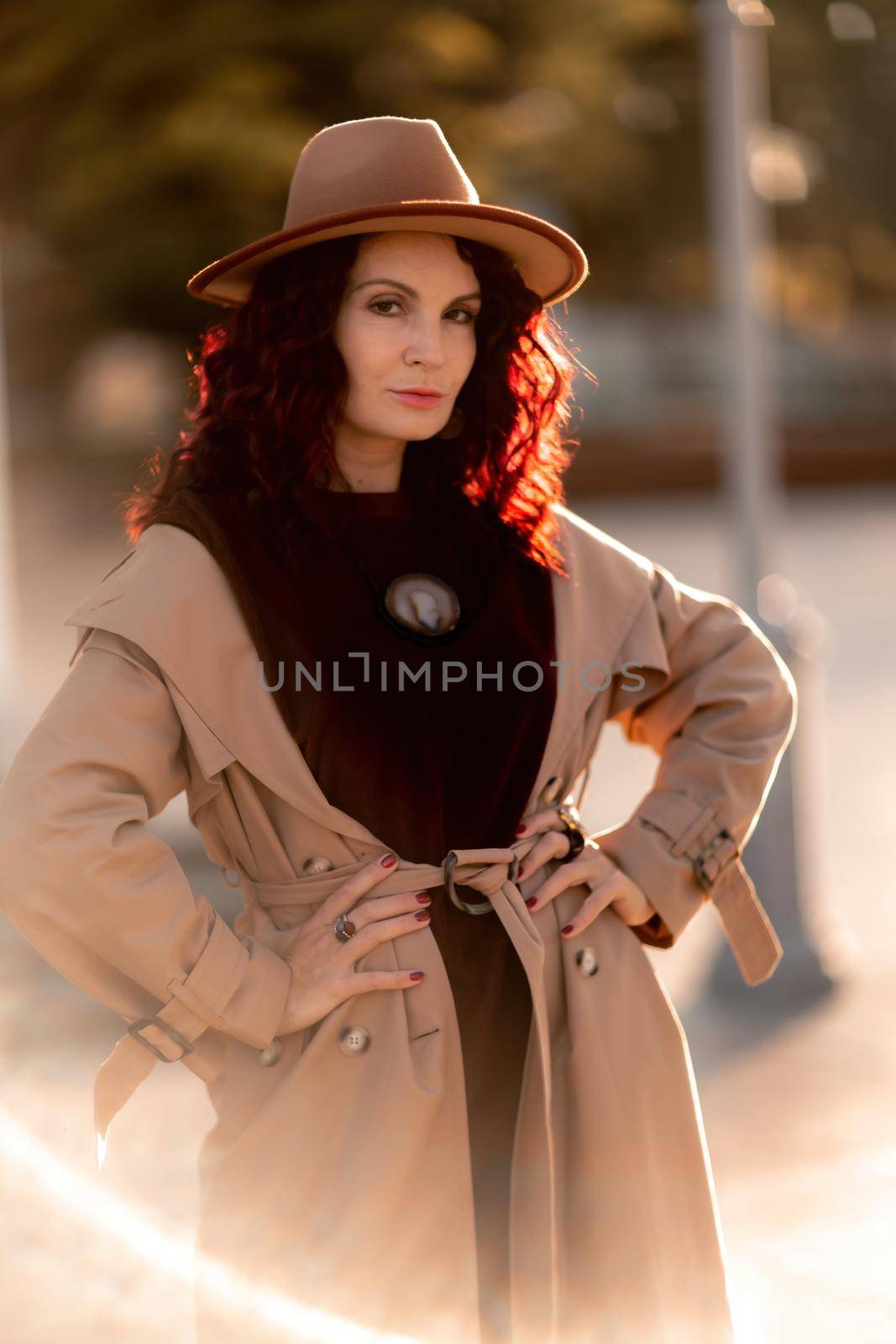 Outdoor fashion portrait of young elegant fashionable brunette woman, model in stylish hat, choker and light raincoat posing at sunset in European city