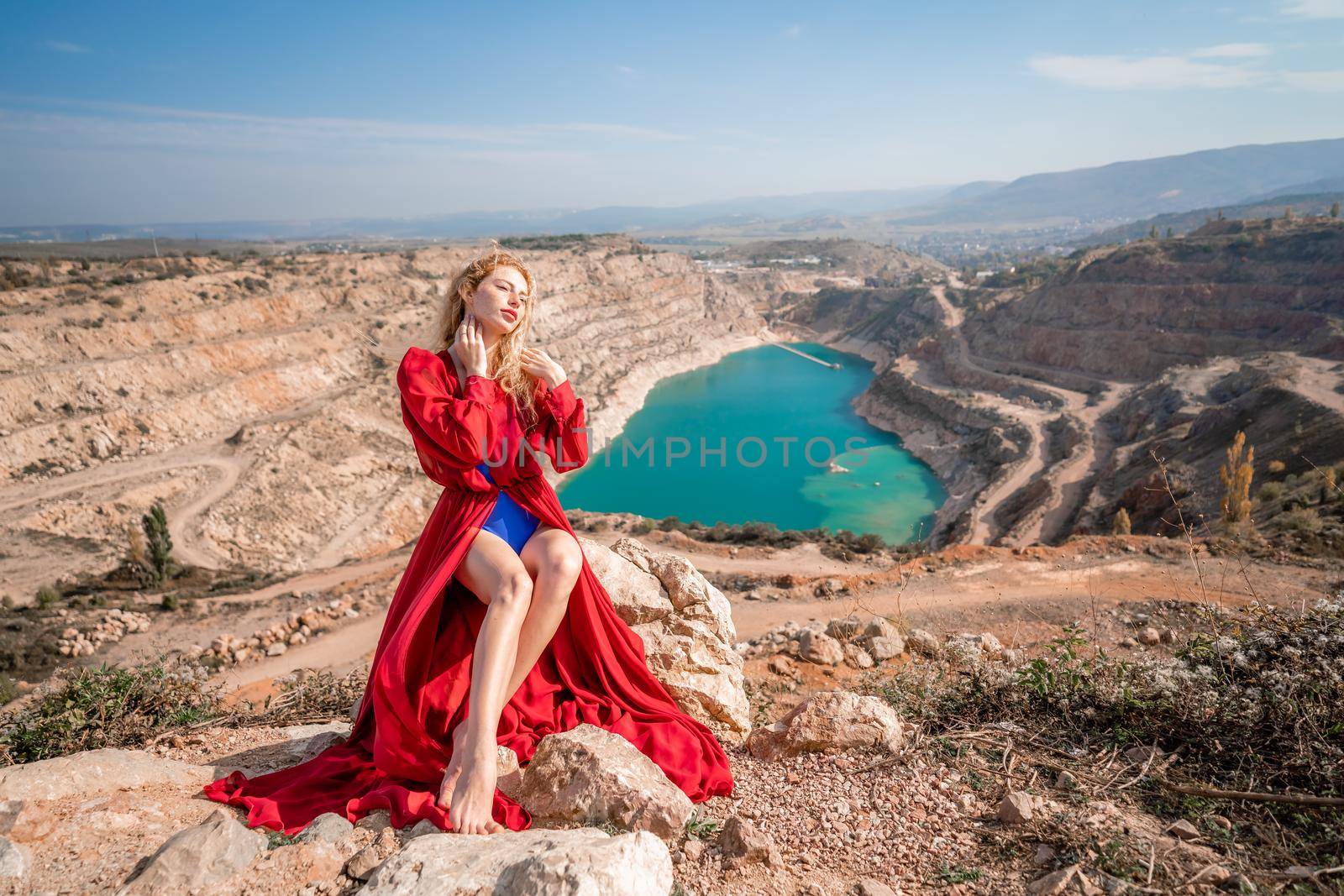 A beautiful girl in a red long dress, Sits on a rock high above the lake in the afternoon. Against the background of the blue sky and the lake in the shape of a heart