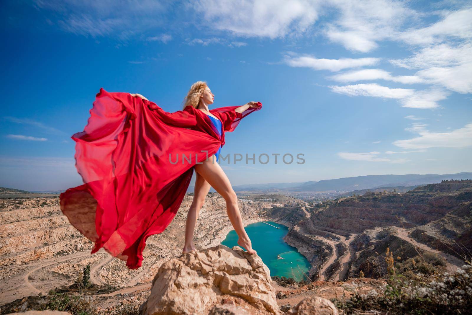 Side view of a beautiful sensual woman in a red long dress posing on a rock high above the lake in the afternoon. Against the background of the blue sky and the lake in the form of a heart by Matiunina