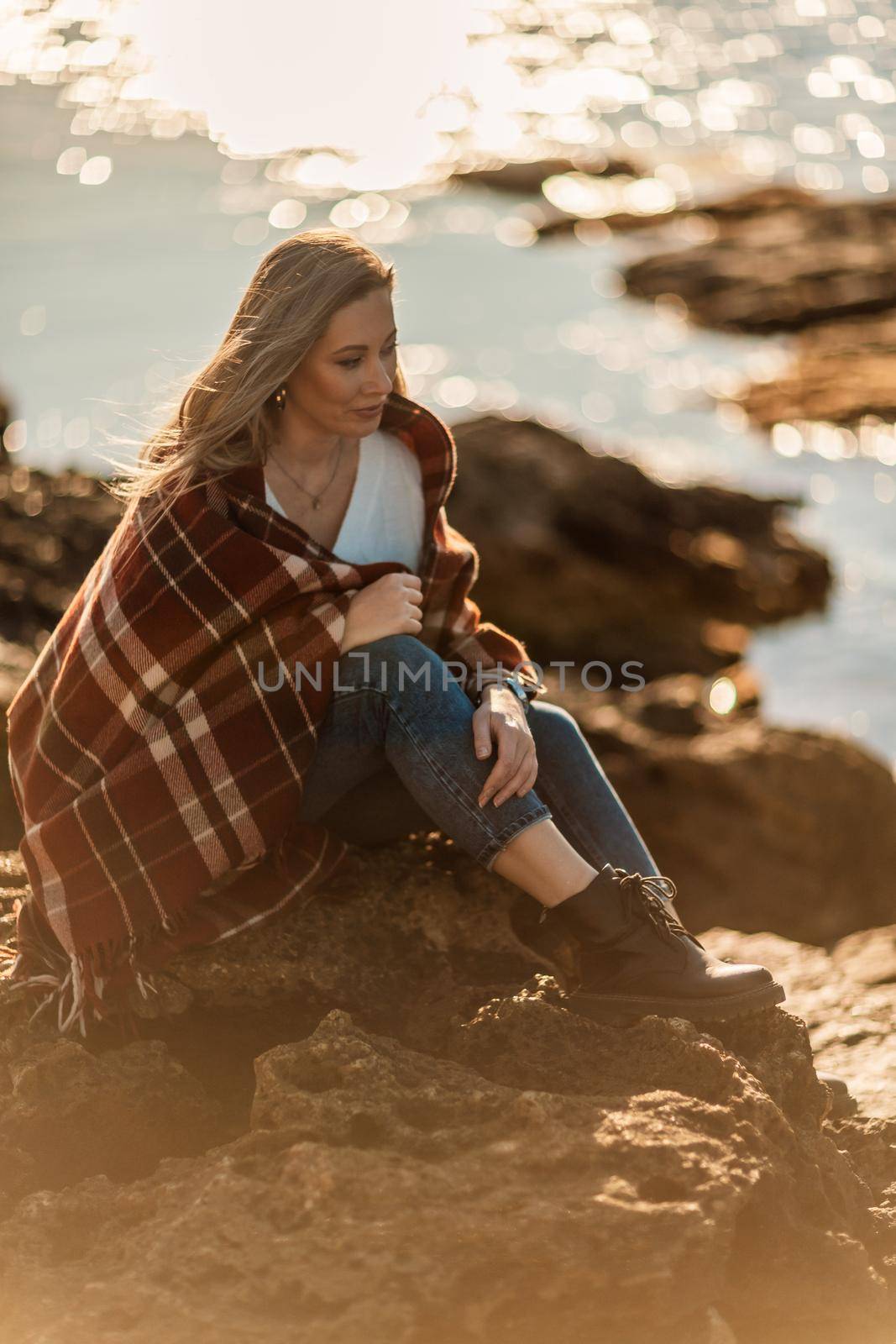 Attractive blonde Caucasian woman enjoying time on the beach at sunset, sitting in a blanket and looking to the side, with the sunset sky and sea in the background. Beach vacation