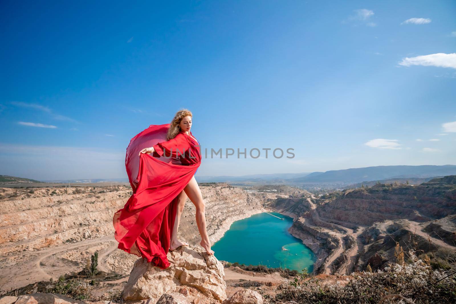 Side view of a beautiful sensual woman in a red long dress posing on a rock high above the lake in the afternoon. Against the background of the blue sky and the lake in the form of a heart.