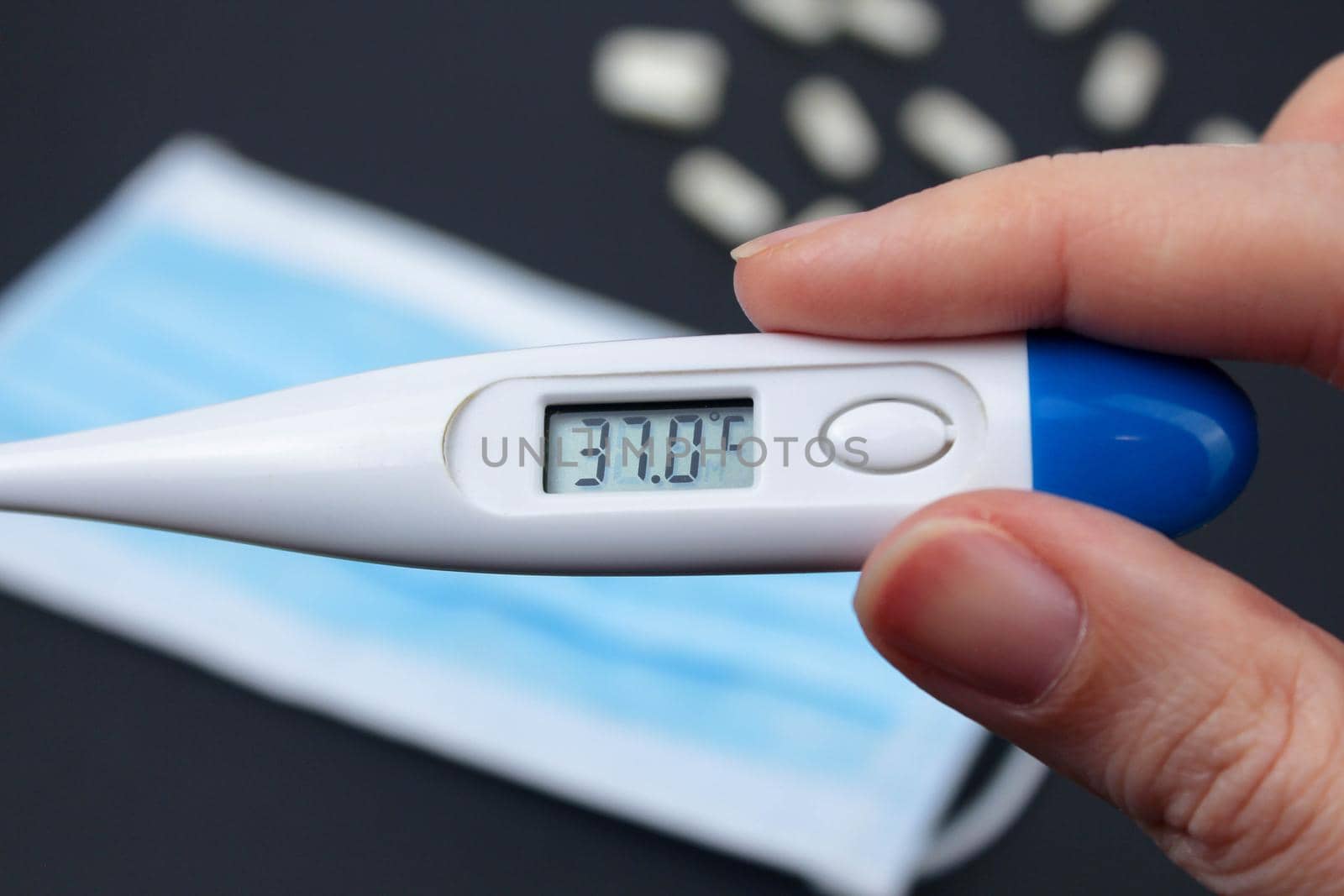 Hand holding white electronic thermometer in the background with packs of tablets and blue medical face mask. Selective focus. by JuliaDorian