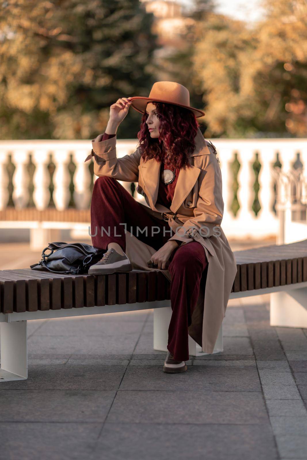 Outdoor fashion portrait of young elegant fashionable brunette woman, model in stylish hat, choker and light raincoat posing at sunset in European city