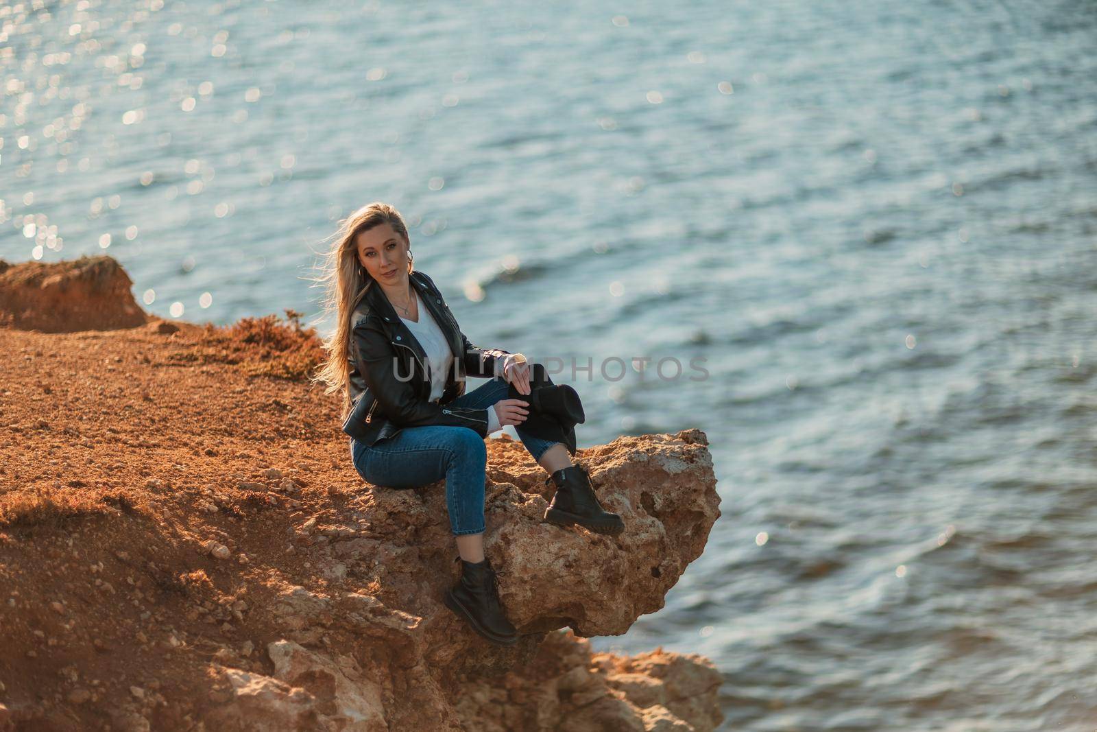 A blonde girl in a stylish black leather jacket is sitting on the seashore