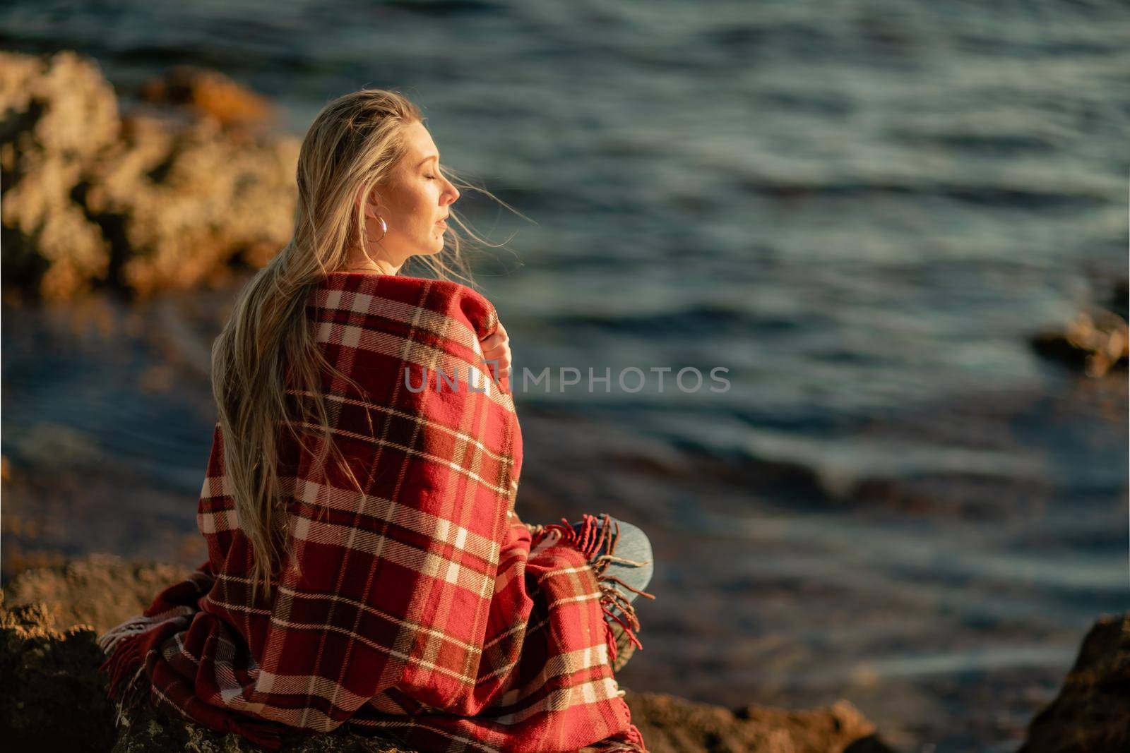 Attractive blonde Caucasian woman enjoying time on the beach at sunset, sitting in a blanket and looking to the side, with the sunset sky and sea in the background. Beach vacation