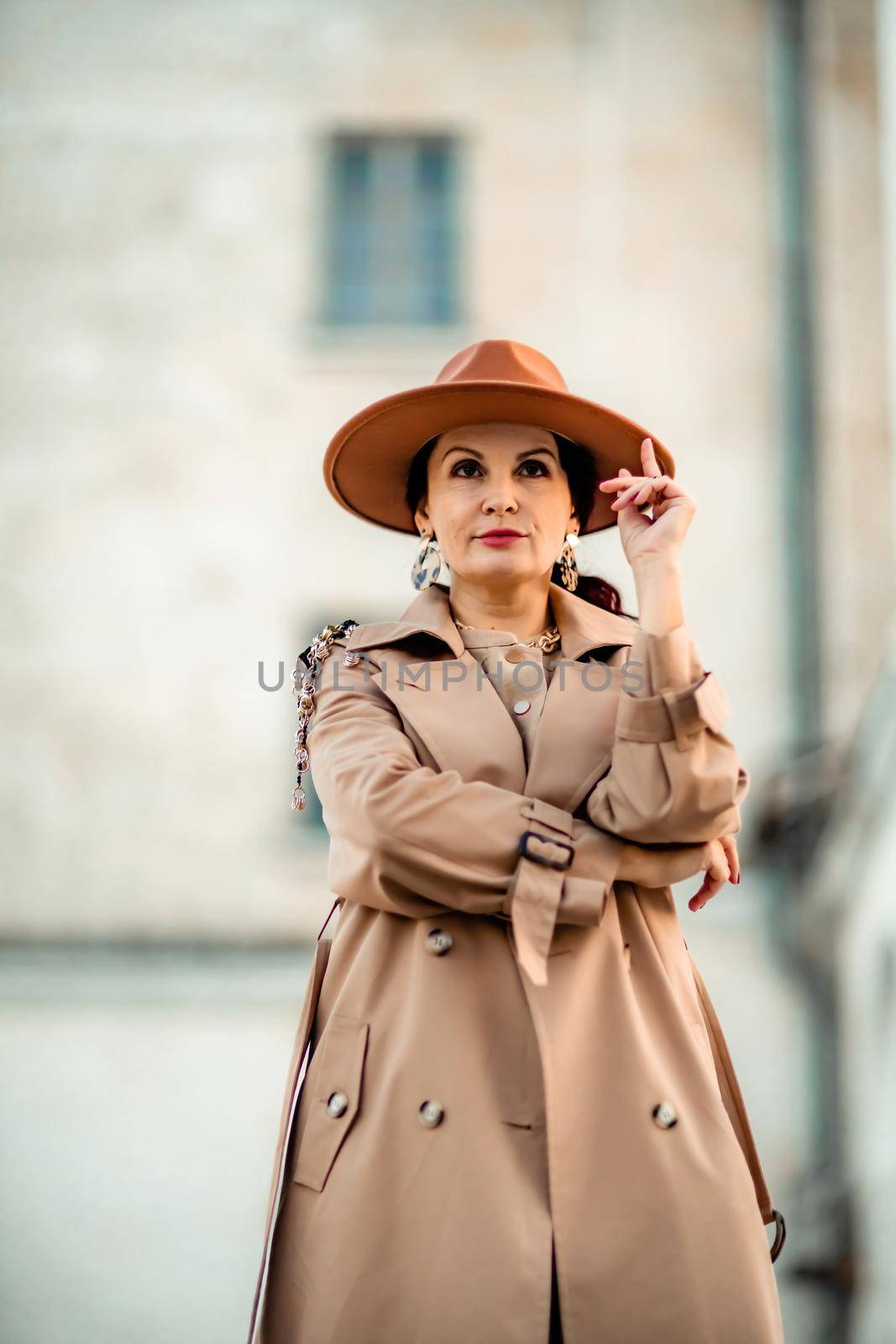 Outdoor fashion portrait of young elegant fashionable brunette woman, model in stylish hat, choker and light raincoat posing at sunset in European city