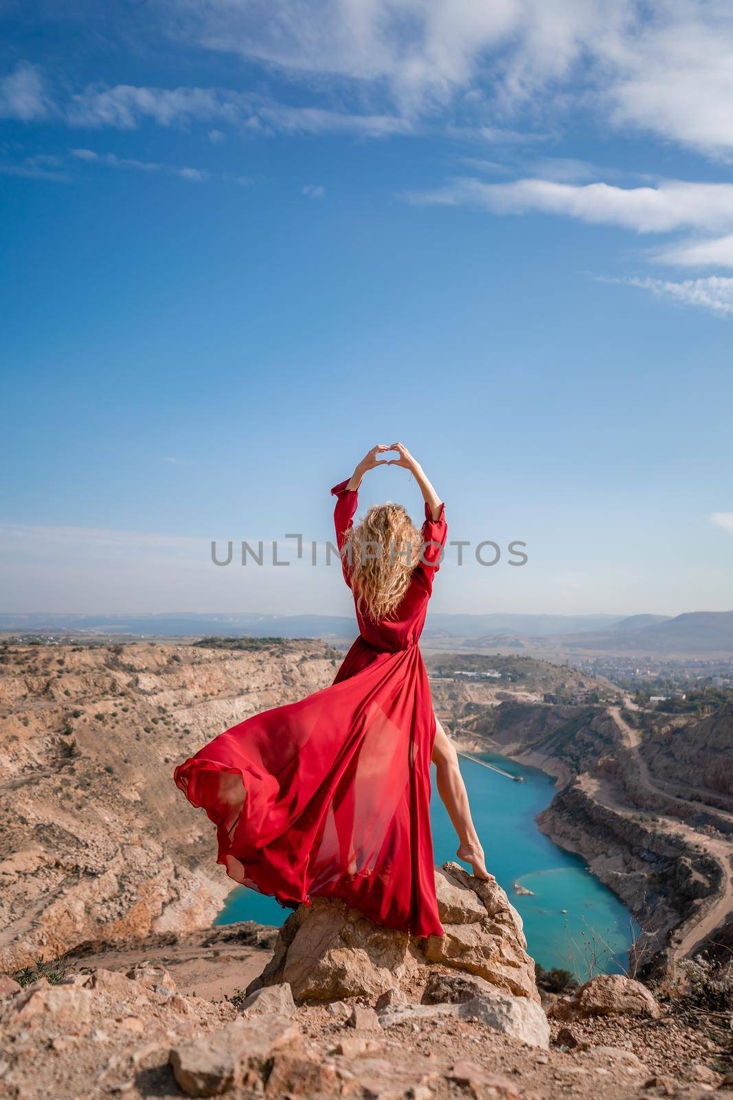 Rear view of a beautiful sensual woman in a red long dress posing on a rock high above the lake in the afternoon. Against the background of the blue sky and the lake in the shape of a heart by Matiunina